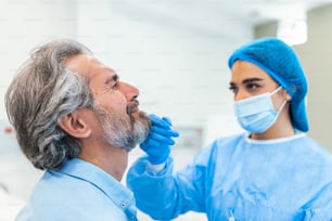 Close up of general practitioner performs coronavirus swab PCR test while wearing face protective mask during covid-19 pandemic. Young woman nurse with surgical mask taking Nose swab for Covid-19