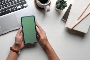 Overhead shot of woman hands holding mock up mobile phone with blank screen at her office desk.