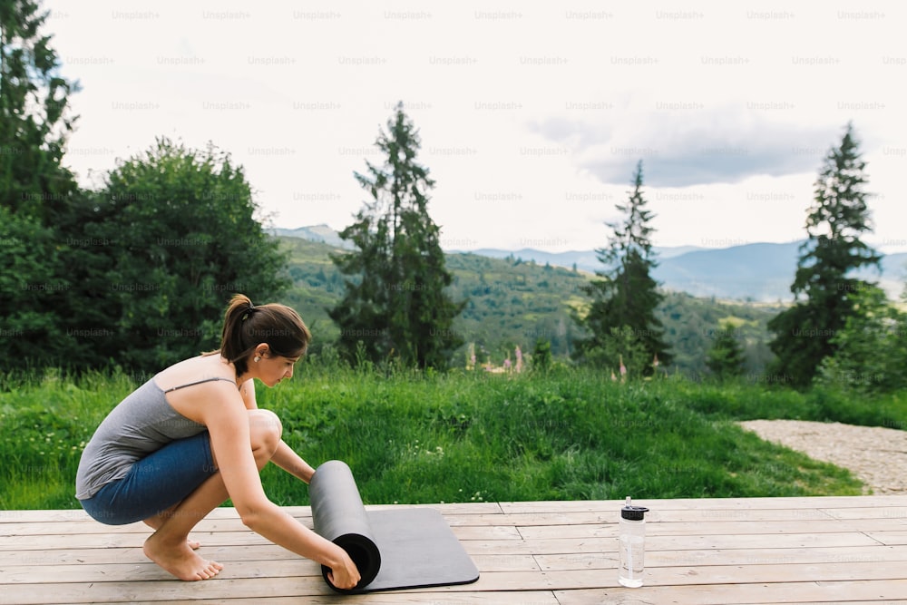 Young fit woman holding yoga mat for exercise on background of sunny mountains hills. Outdoor workout and training. Casual female standing on wooden terrace among trees