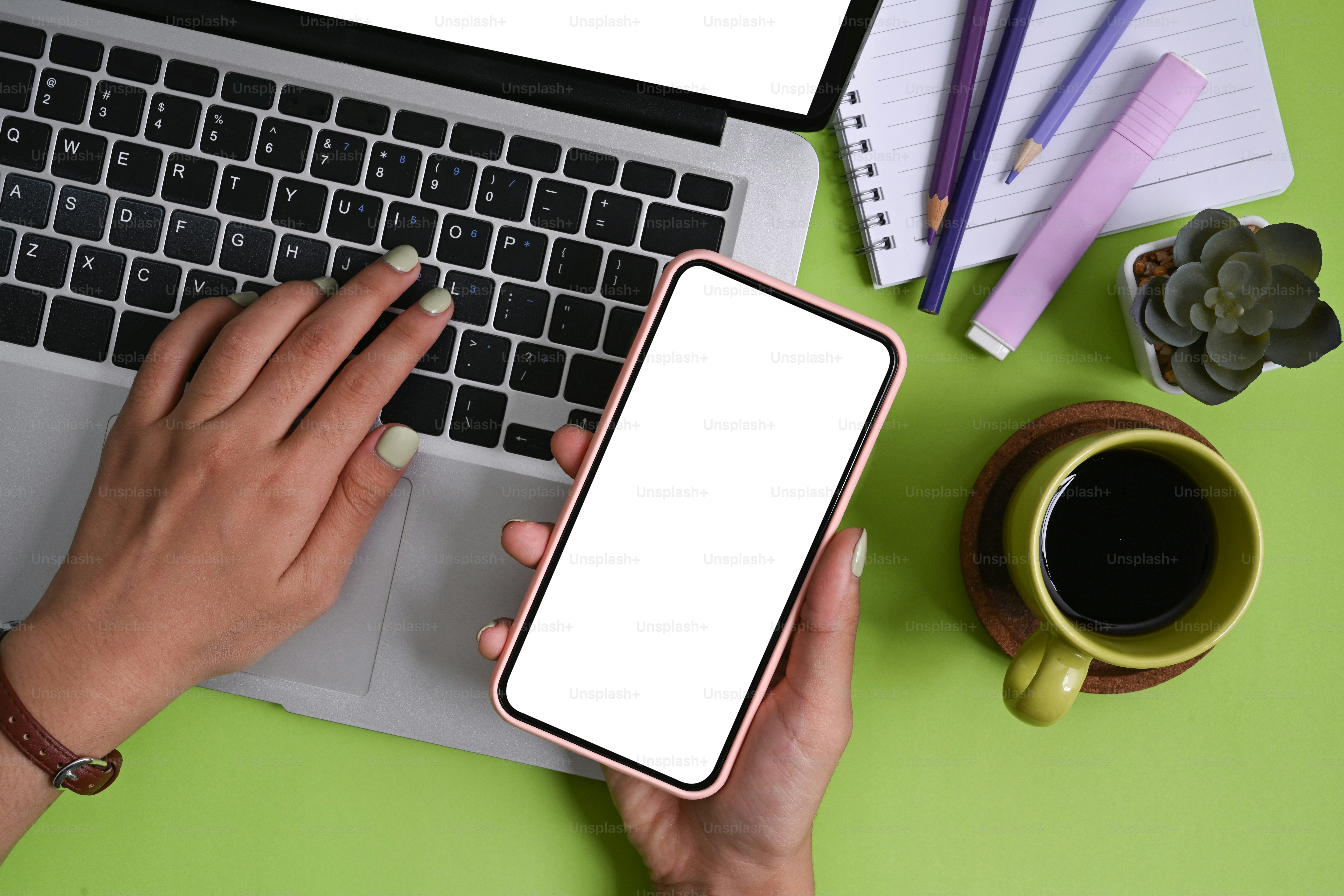 Overhead view of woman holding smart phone and using laptop computer on green background.
