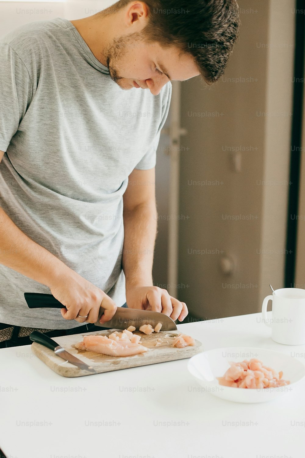 Joven cortando filete de pollo con cuchillo en tabla de madera en cocina blanca moderna. Proceso de cocción y preparación de la carne, con las manos en alto. Concepto de cocina casera