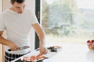 Young man putting tomato slices on bread and making avocado toasts in modern white kitchen. Process of making toasts. Healthy eating and Home cooking concept