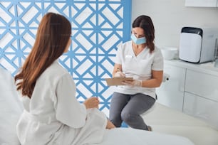 Nice female cosmetologist in medical mask sitting on chair and writing on clipboard while listening to patient