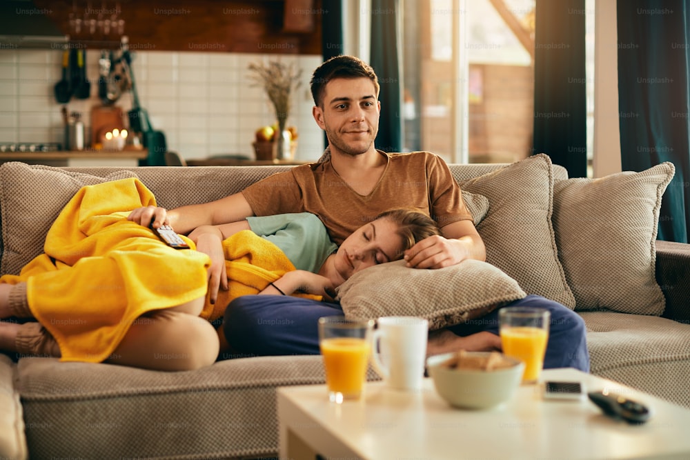Jeune couple se relaxant dans le salon. Un homme souriant regarde la télévision tandis qu’une femme dort sur le canapé.