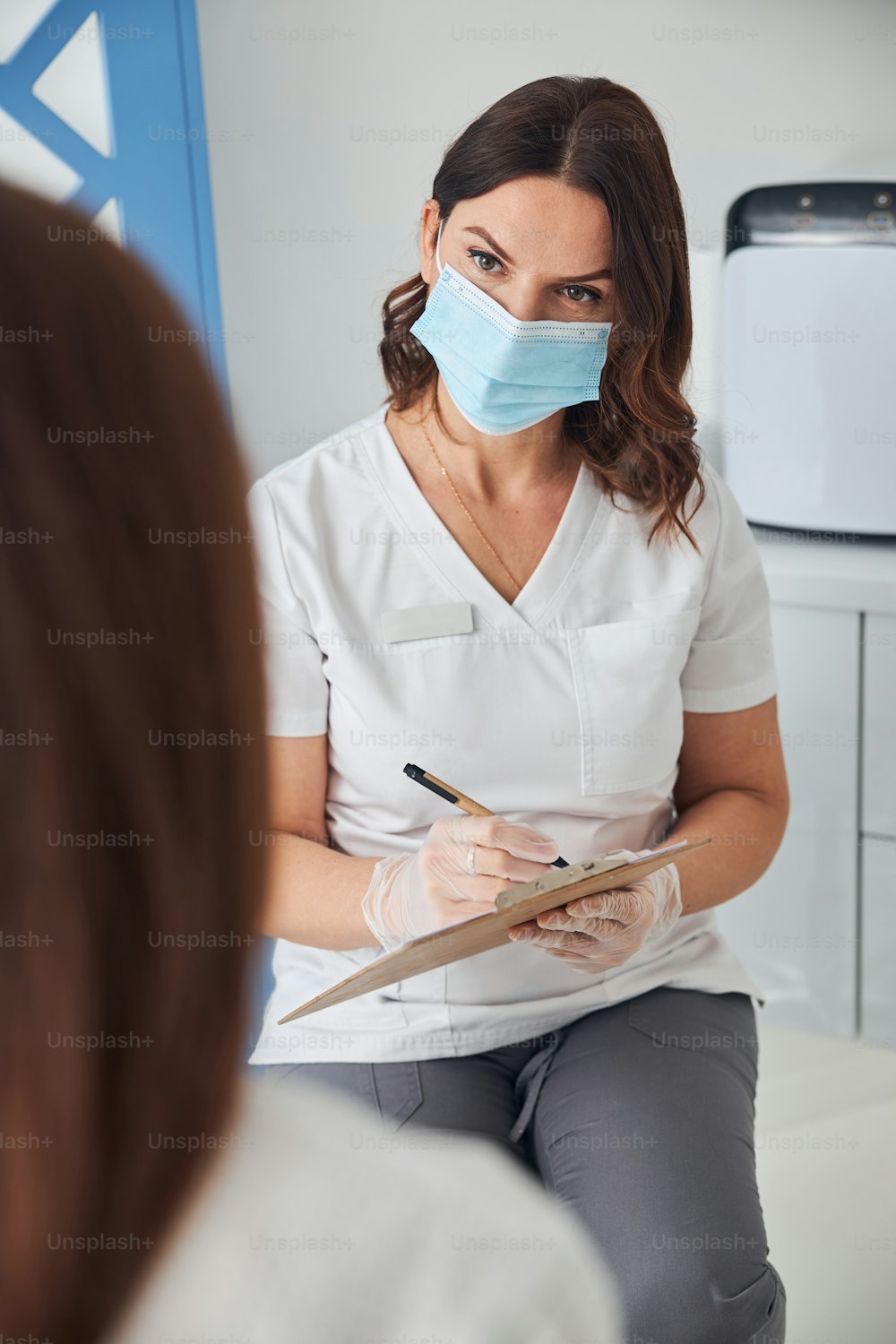 Charming woman cosmetologist wearing medical face mask while talking with client and writing on clipboard