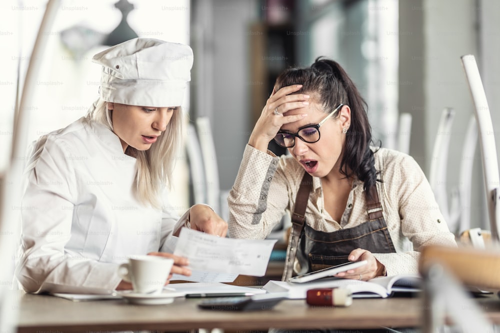 Shock over bookkeeping by female chef and restaurant owner, sitting over paperwork.