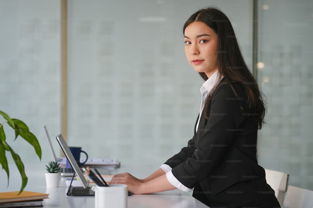 Side view of confident businesswoman in suit sitting in modern workplace and looking at camera.