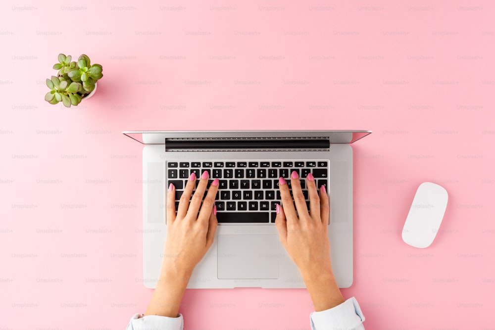 컴퓨터 마우스와 꽃이 있는 분홍색 배경에 노트북을 사용하는 여성 손. 사무실 데스크톱. 상위 뷰