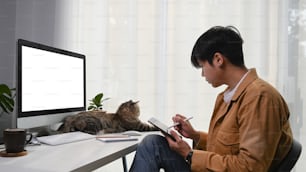 デジタルタブレットで作業する若い男性のグラフィックデザイナーと、彼の前に横たわっているかわいい猫の側面図。