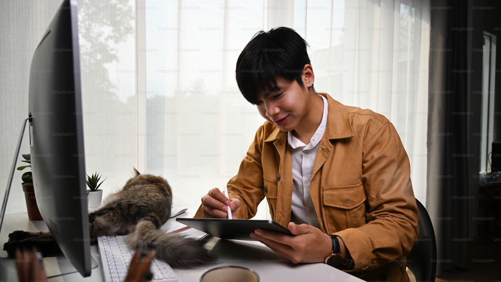 Joven sonriente diseñador gráfico que trabaja en línea con una tableta digital y su gato acostado frente a él.