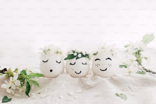 Natural easter eggs with drawn cute faces in floral wreaths on linen fabric with blooming spring branch, petals and green leaves in soft light. Happy Easter! Space for text. Eco friendly holiday
