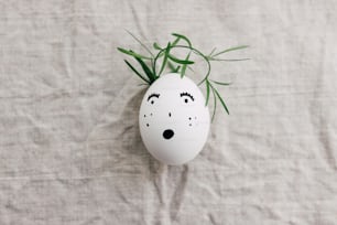 Natural egg with drawn surprised face in cute floral crown on linen fabric. Happy Easter! Eco friendly zero waste holiday concept. Top view with space for text