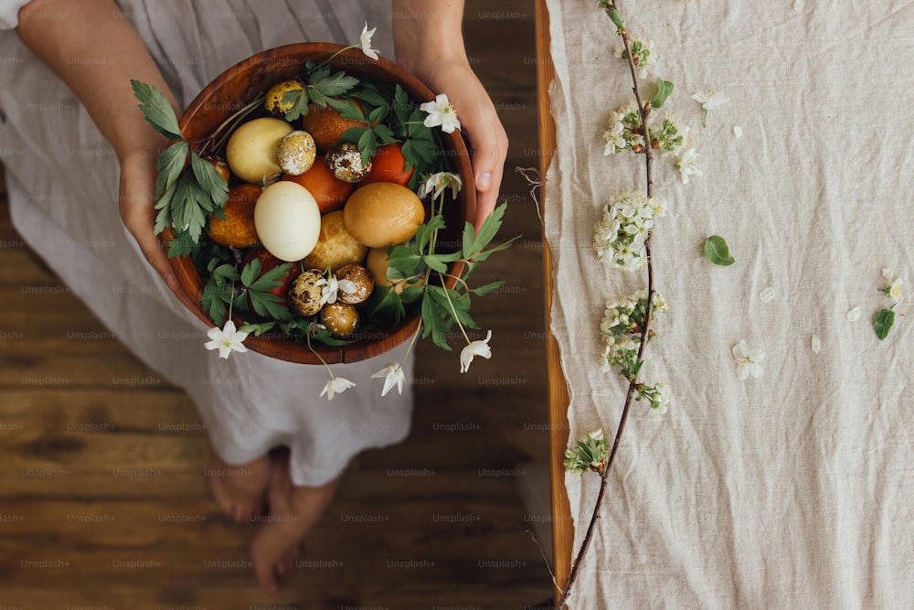 Easter eggs and spring flowers in wooden bowl in hands on background of woman in rustic linen dress and table. Stylish easter and quail eggs in natural dye and spring blooms. Aesthetic holiday