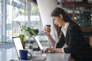 Vista lateral de una atractiva mujer de negocios que sostiene una taza de café y trabaja en una computadora portátil en una oficina moderna.