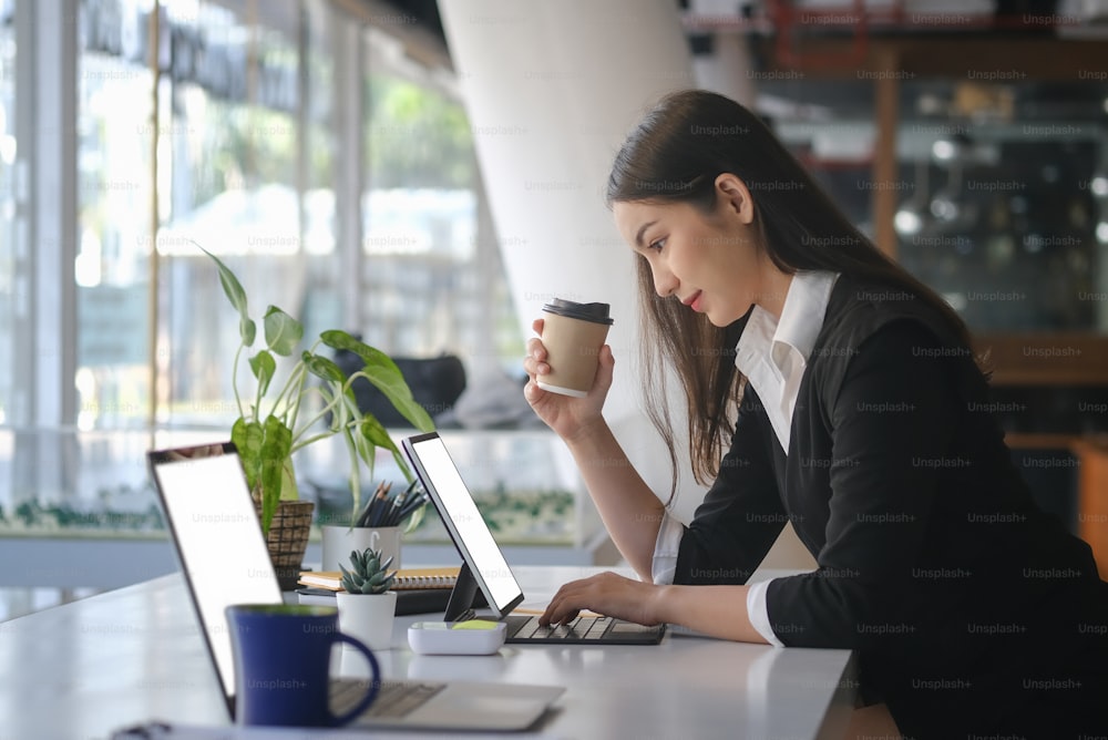 Vue latérale d’une femme d’affaires attrayante tenant une tasse de café et travaillant sur un ordinateur portable dans un bureau moderne.