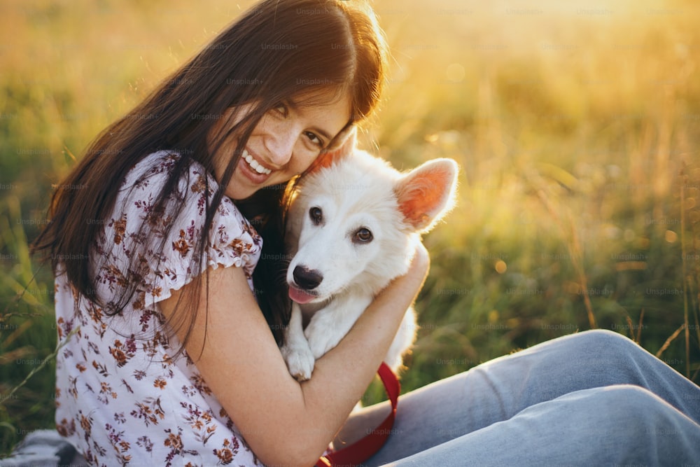 따뜻한 일몰 빛 아래 여름 초원에서 귀여운 하얀 강아지와 껴안고 있는 행복한 여자. 사랑스러운 스위스 양치기 강아지와 함께 휴식을 취하는 캐주얼한 젊은 여성. 행복. 애완 동물과 함께하는 여름 휴가