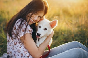 Happy woman cuddling with cute white puppy in summer meadow in warm sunset light. Casual young female relaxing with adorable swiss shepherd puppy. Happiness. Summer vacation with pet