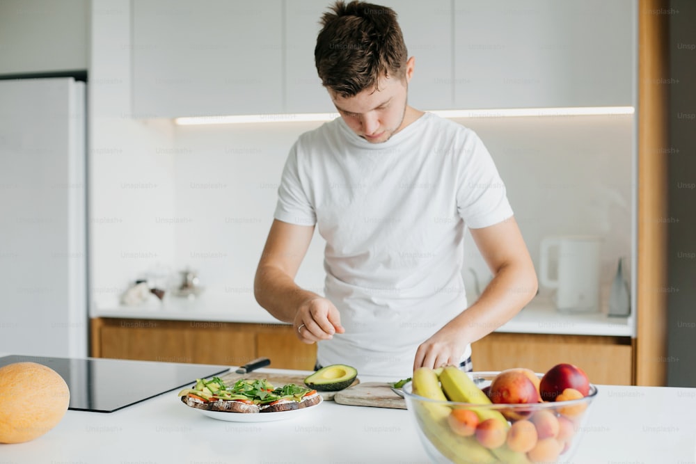 Young man making toasts with avocado, tomato, arugula, cheese in modern white kitchen. Healthy eating and Home cooking concept. Hands putting arugula on sandwich