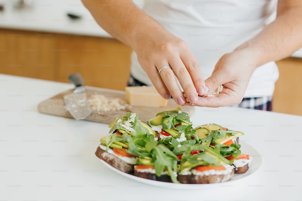 현대적인 흰색 주방에서 아보카도, 토마토, 아루굴라, 치즈로 토스트를 만드는 청년. 건강한 식생활과 가정 요리 개념. 샌드위치에 집에서 만든 파마산 치즈를 넣는 손