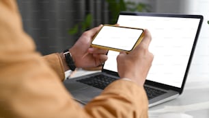 Close up view of man hands holding horizontal mobile phone while sitting in front of his computer tablet at home office.