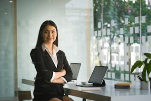 Confident businesswoman in suit sitting with arms crossed  and smiling to camera in modern office.