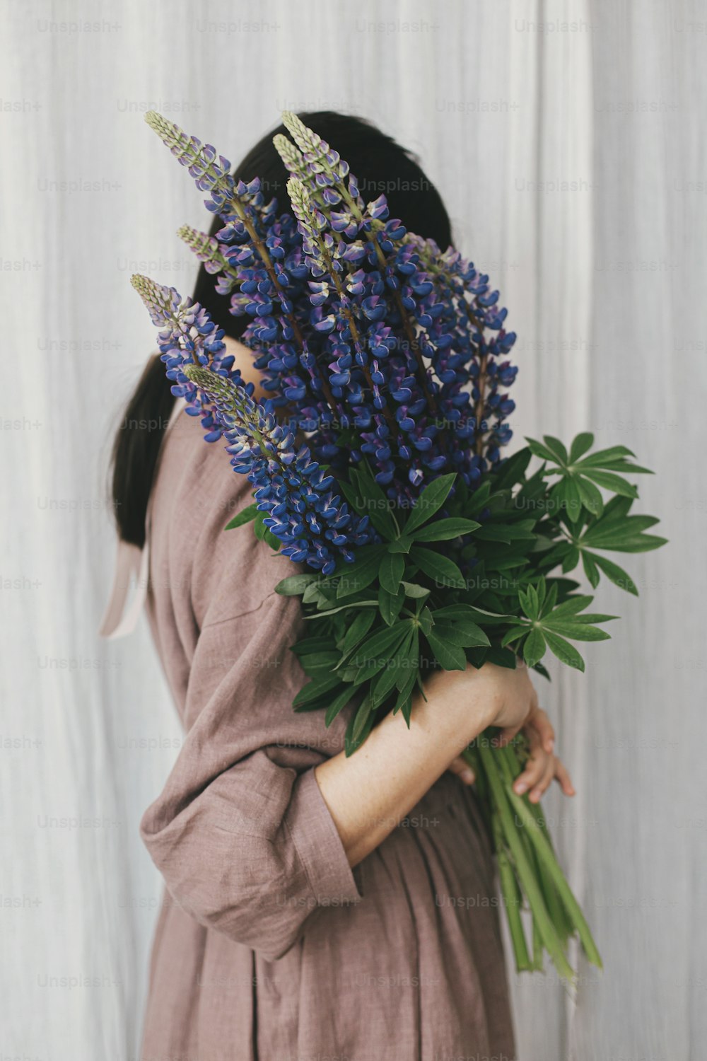 Stylish woman in linen dress holding lupine bouquet on background of pastel fabric. Simple slow living. Young female in boho rustic dress hiding face with lupine wildflowers. Aesthetics