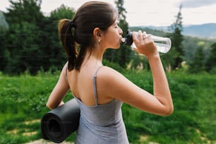 Young woman drinking water after training on background of sunny mountain hills. Outdoor workout. Healthy lifestyle. Sporty casual female holding yoga mat and drinking water from bottle among trees