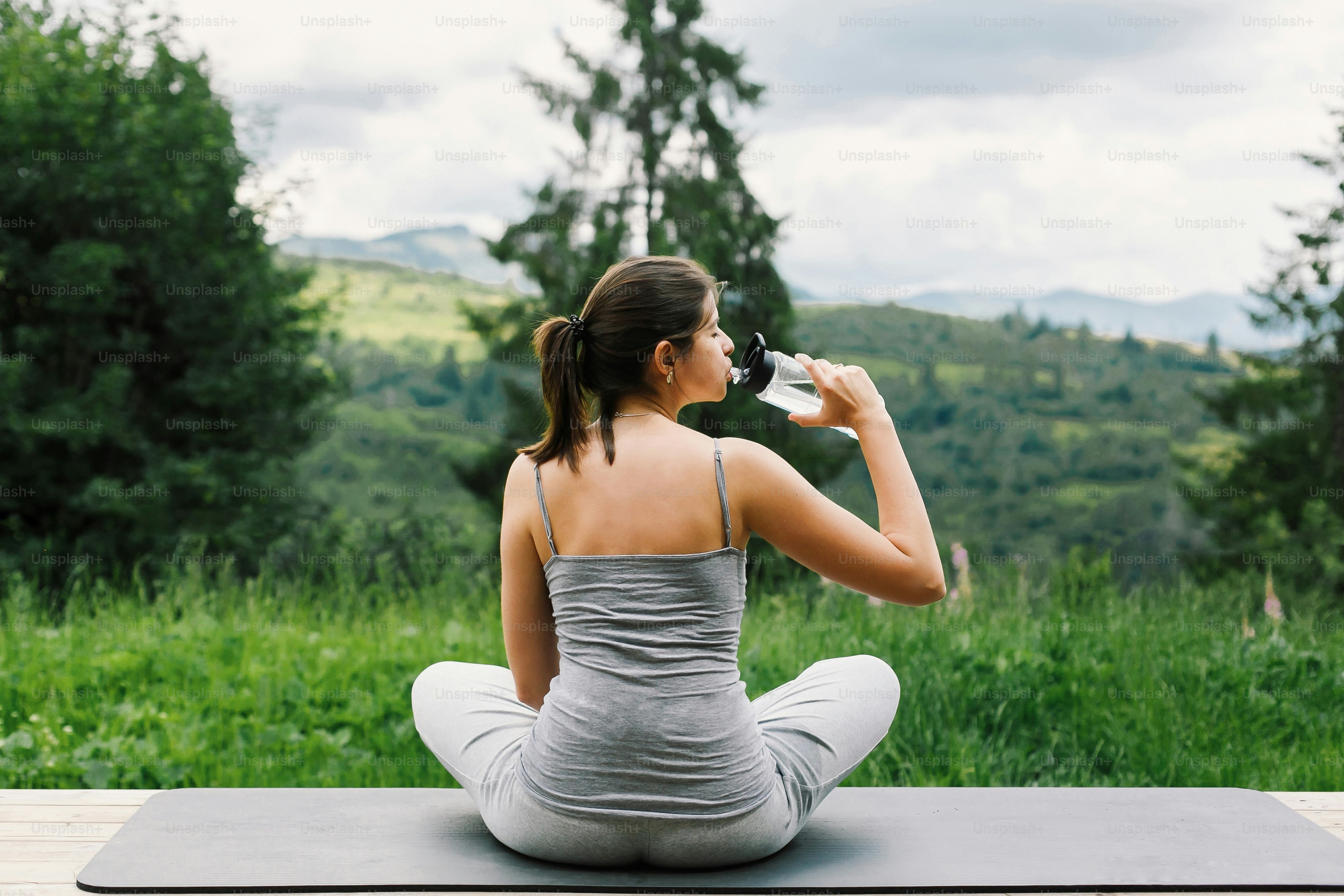 Young fit woman drinking water after practising yoga or training on background of sunny mountains hills. Outdoor workout. Healthy lifestyle. Sporty Female relaxing on mat among trees