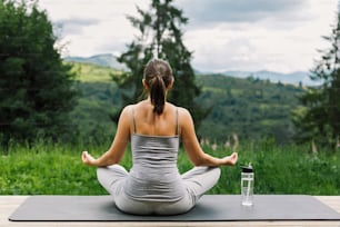 Young fit woman practising meditation on yoga mat on background of sunny mountains hills. Outdoor workout and training. Healthy lifestyle. Sporty Female doing yoga on wooden terrace among trees