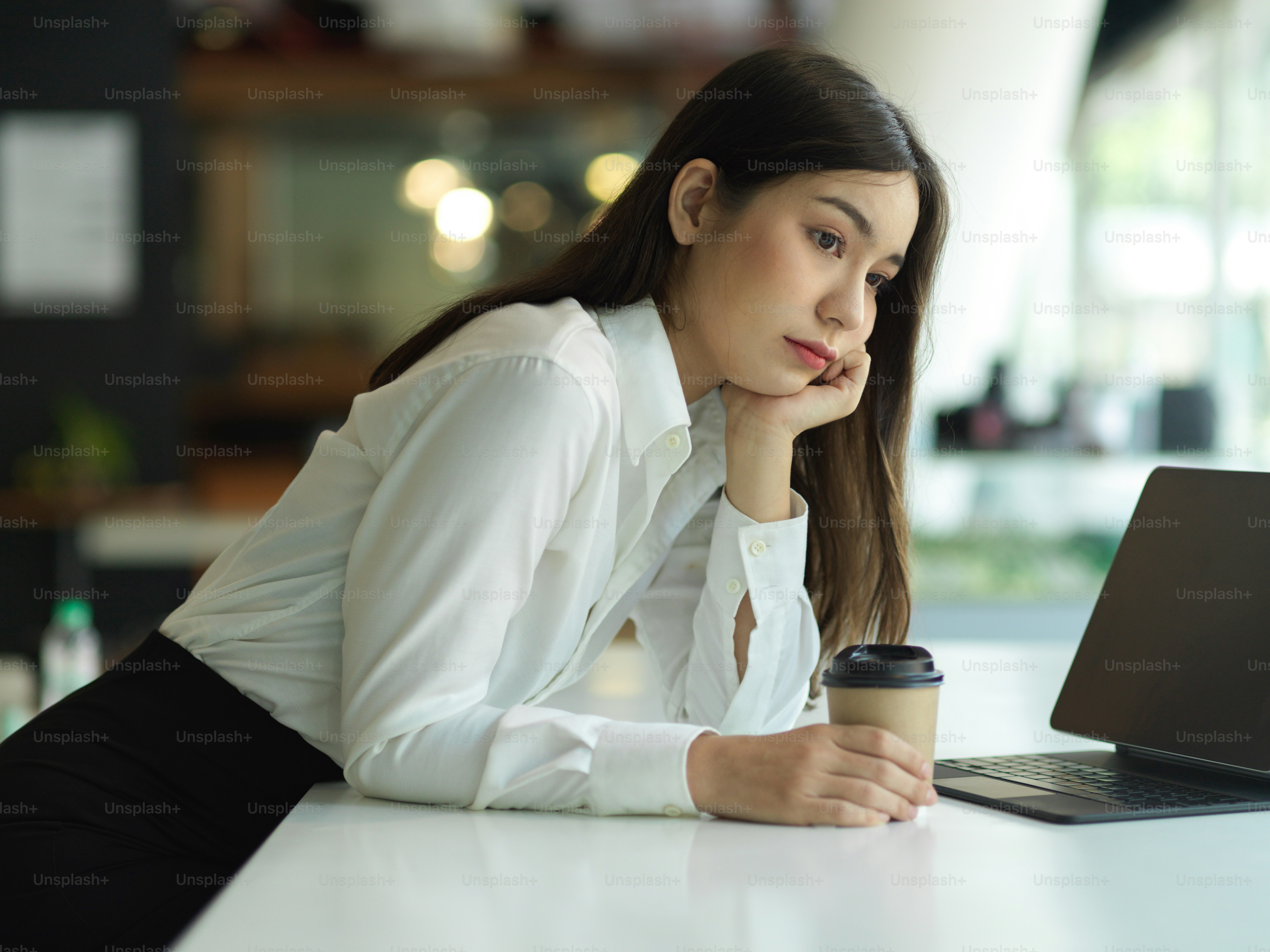 頬に手を当てて紙コップを持ち、事務室で仕事のことを考えている女性労働者の側面図