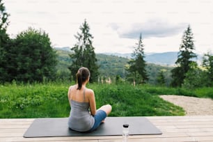 Young fit woman relaxing or practising meditation on yoga mat on background of sunny mountains hills. Outdoor workout and training. Healthy lifestyle. Sporty Female doing yoga on wooden terrace