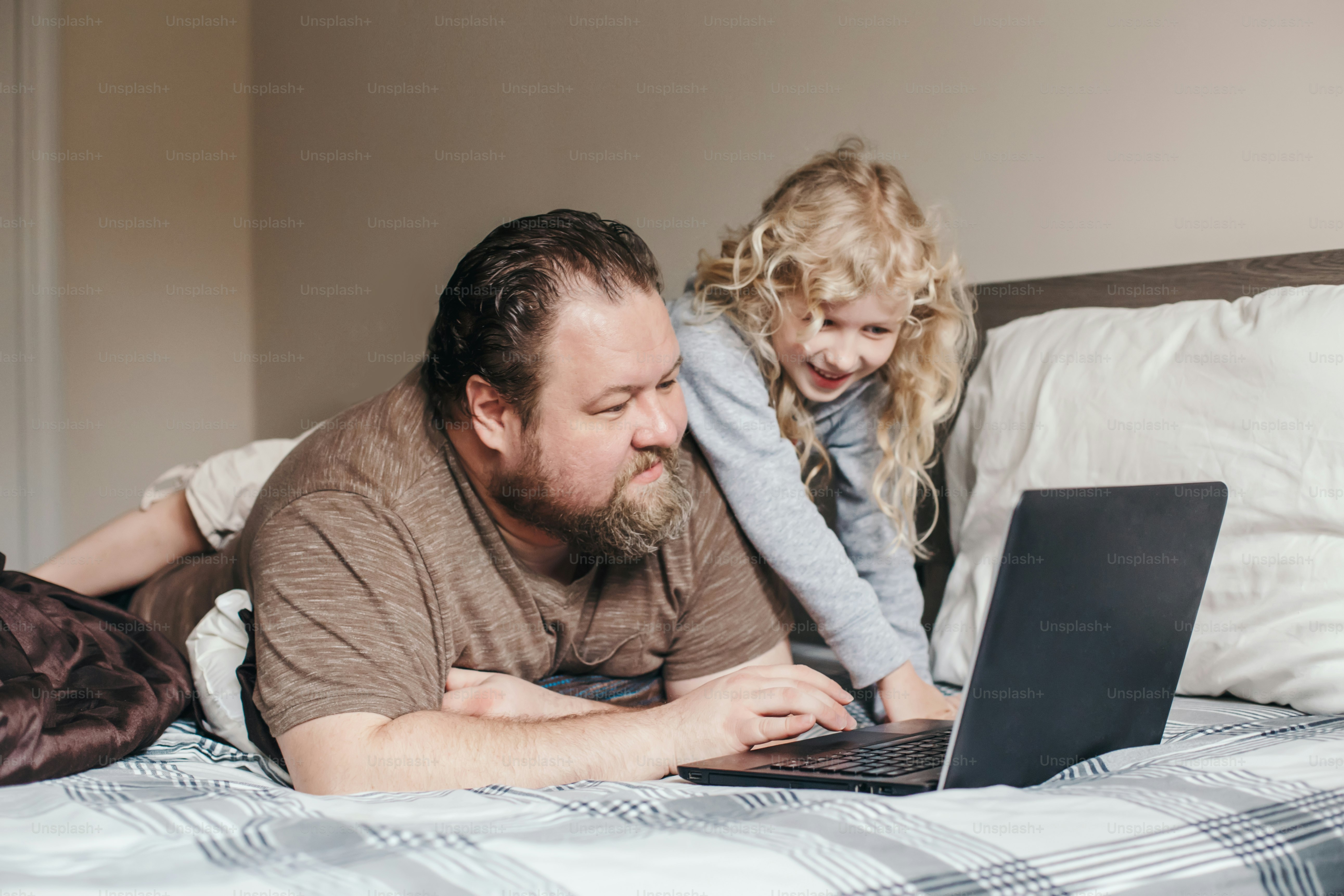 Trabaja desde casa con niños. Padre trabajando en una computadora portátil en el dormitorio con una hija pequeña en su espalda. Momento familiar divertido y sincero. Nueva normalidad durante el confinamiento por la cuarentena por el coronavirus.