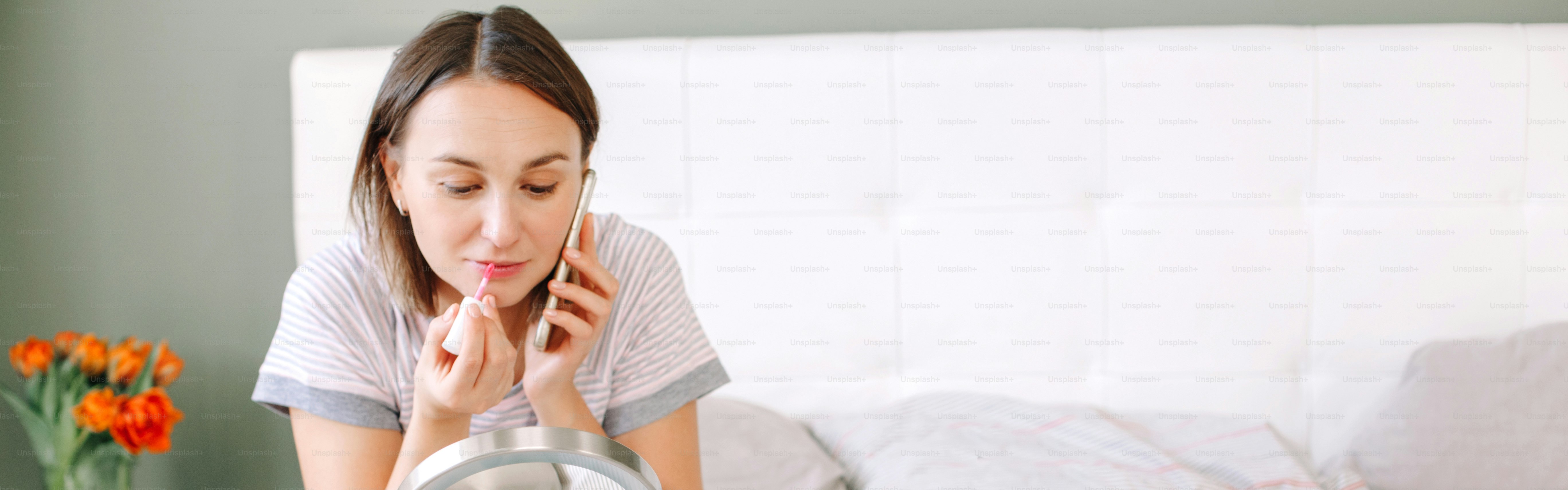 Middle age Caucasian woman applying makeup decorative cosmetics and talking on phone. Woman sitting in bed at home doing morning routine and chatting with friend. Web banner header.