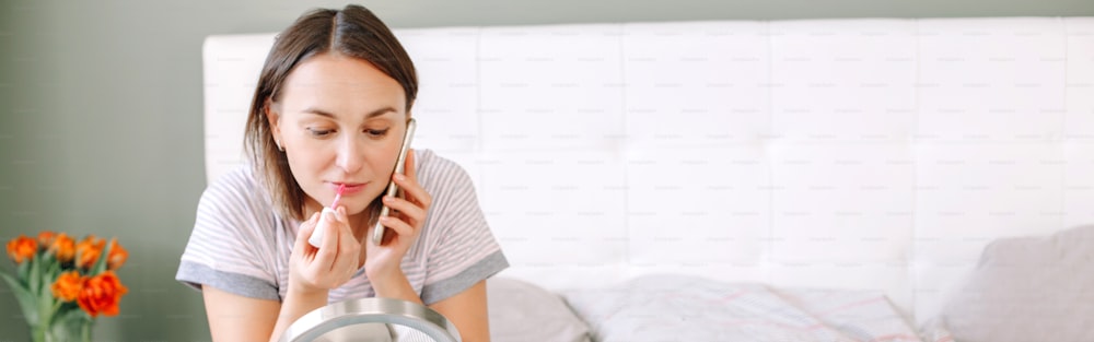 Mujer caucásica de mediana edad que se maquilla, decora, cosméticos y habla por teléfono. Mujer sentada en la cama en casa haciendo la rutina matutina y charlando con un amigo. Encabezado de banner web.