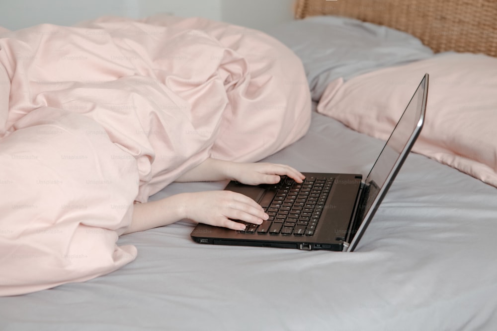 Gelangweiltes lustiges Kind, das unter einer Decke im Bett liegt und in der virtuellen Online-Schulklasse lernt. Hände tippen auf der Computertastatur. Kind, das zu Hause am Laptop-Internet arbeitet. Homeschooling für Kinder.