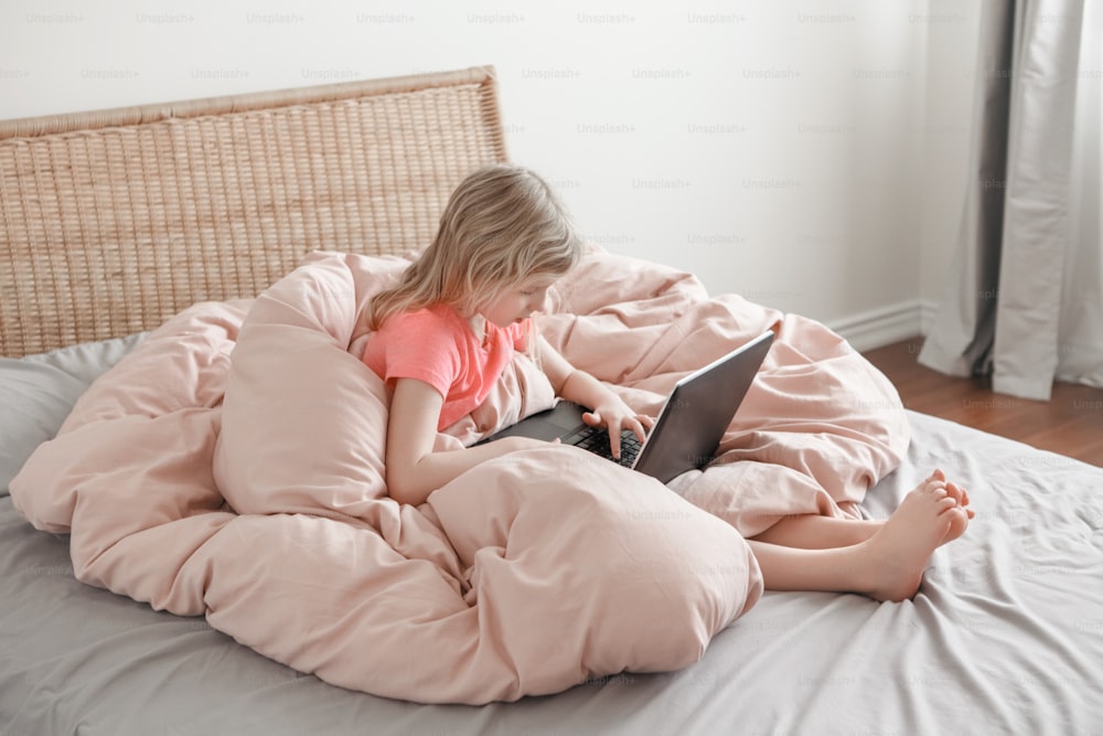 Girl sitting in bed and learning in virtual online school class. Kid typing working on laptop Internet at home. Child using wireless technology education. Homeschooling for children.