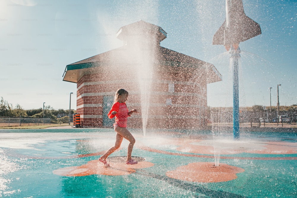 Cute adorable Caucasian funny girl playing on splash pad playground on summer day. Happy child having fun in water. Seasonal water sport recreational activity for kids outdoor.