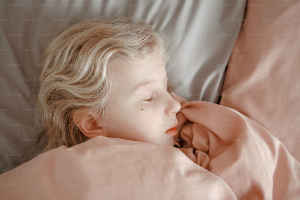 Primo piano di una ragazza bionda caucasica carina che dorme sognando a letto a casa. Bambino stanco del sonno con gli occhi chiusi sotto la coperta rosa in camera da letto. Routine mattutina. Sogni d'oro.