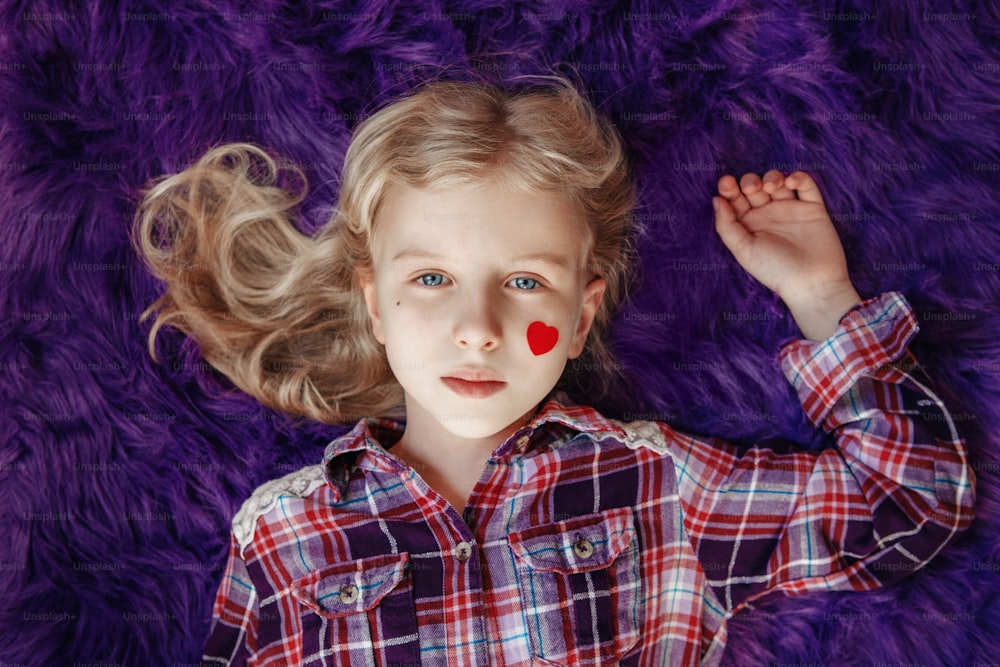 Happy Valentine Day holiday. Cute pretty blonde Caucasian girl with small red paper heart on her cheek lying on purple fluffy rug carpet at home. Child kid celebrating love day. View from above.