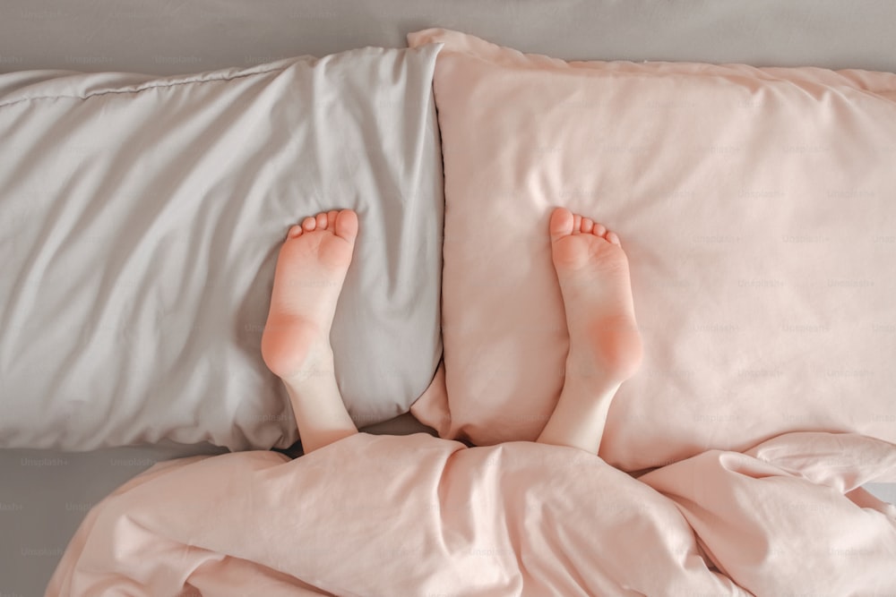 子供の足、家のベッドの枕に横たわっている足。毛布の下でかくれんぼをする子供。ピンクのかわいい愛らしい赤ちゃんのかかとがベッドで逆さまになりました。幸せな本物の子供時代のライフスタイル。