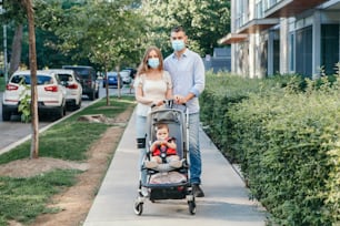 Madre e padre caucasici in maschere per il viso che camminano con il bambino nel passeggino. Famiglia che passeggia insieme all'aperto sulla strada della città il giorno d'estate. Nuova normalità nella pandemia di coronavirus covid-19.
