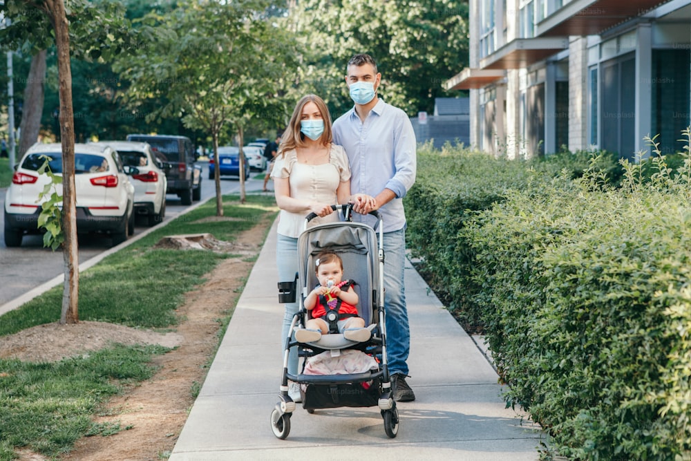 Caucasian mother and father in face masks walking with baby in stroller. Family strolling together outdoor on city street on summer day. New normal in coronavirus covid-19 pandemic.