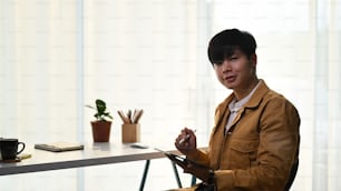 Joven asiático sosteniendo una tableta digital y sonriendo a la cámara mientras está sentado en la oficina de su casa.
