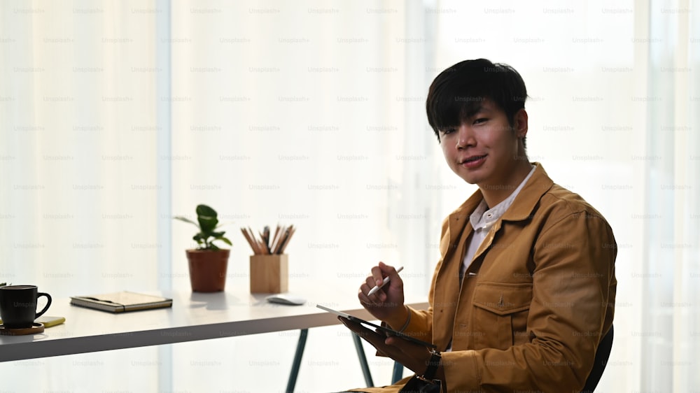 Young asian man holding digital tablet and smiling to camera while sitting in his home office.