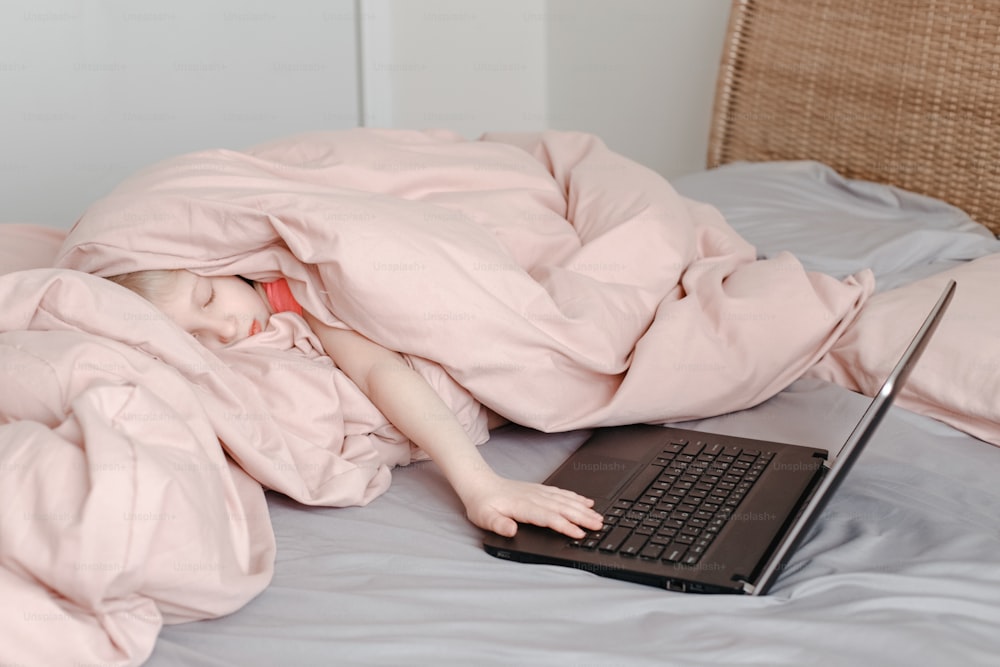 Menina sonolenta e cansada deitada na cama sob o cobertor e aprendendo na aula virtual da escola on-line. Criança trabalhando na Internet do laptop em casa. Criança usando educação de tecnologia sem fio. Homeschooling para crianças.