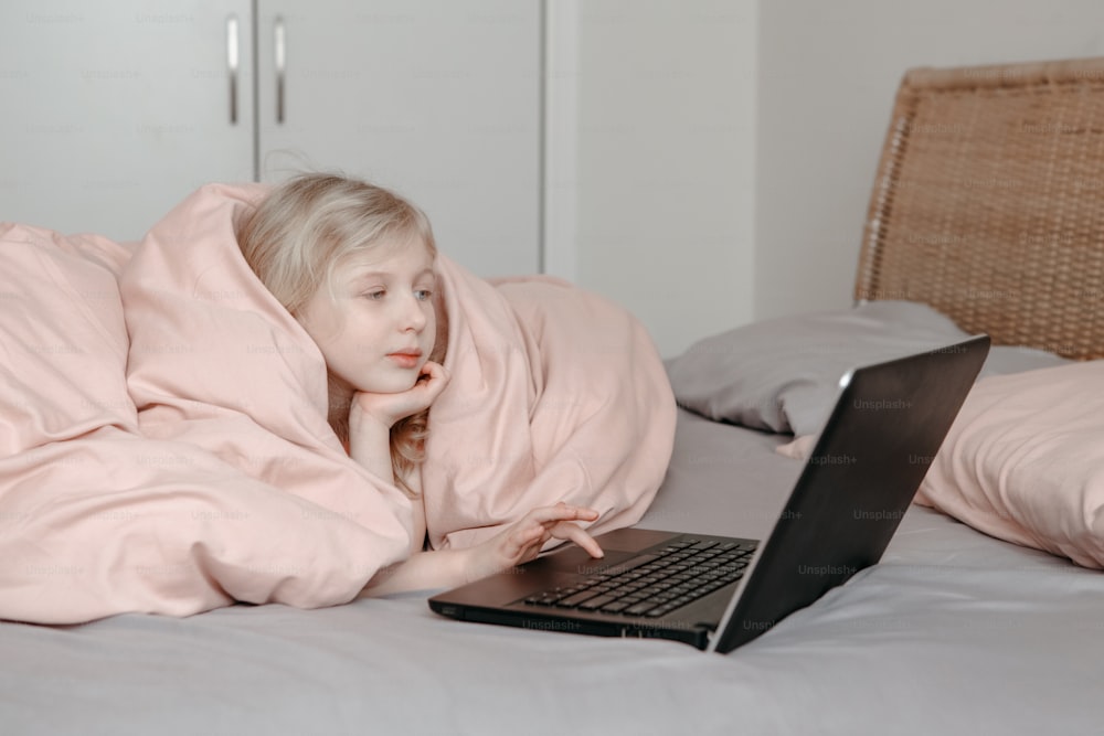 Menina entediada deitada na cama debaixo do cobertor e aprendendo na aula virtual da escola on-line. Criança trabalhando na Internet do laptop em casa. Criança usando educação de tecnologia sem fio. Homeschooling para crianças.