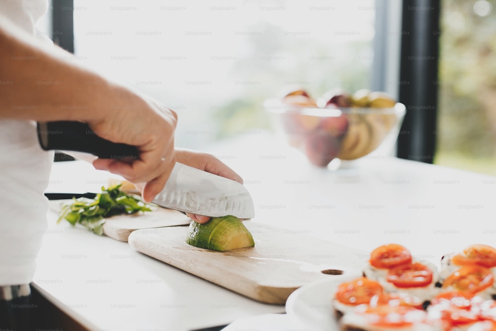 Young man cutting avocado and making toasts with tomato, arugula, cheese in modern white kitchen. Healthy eating and Home cooking concept. Perfectly ripe avocado in hands