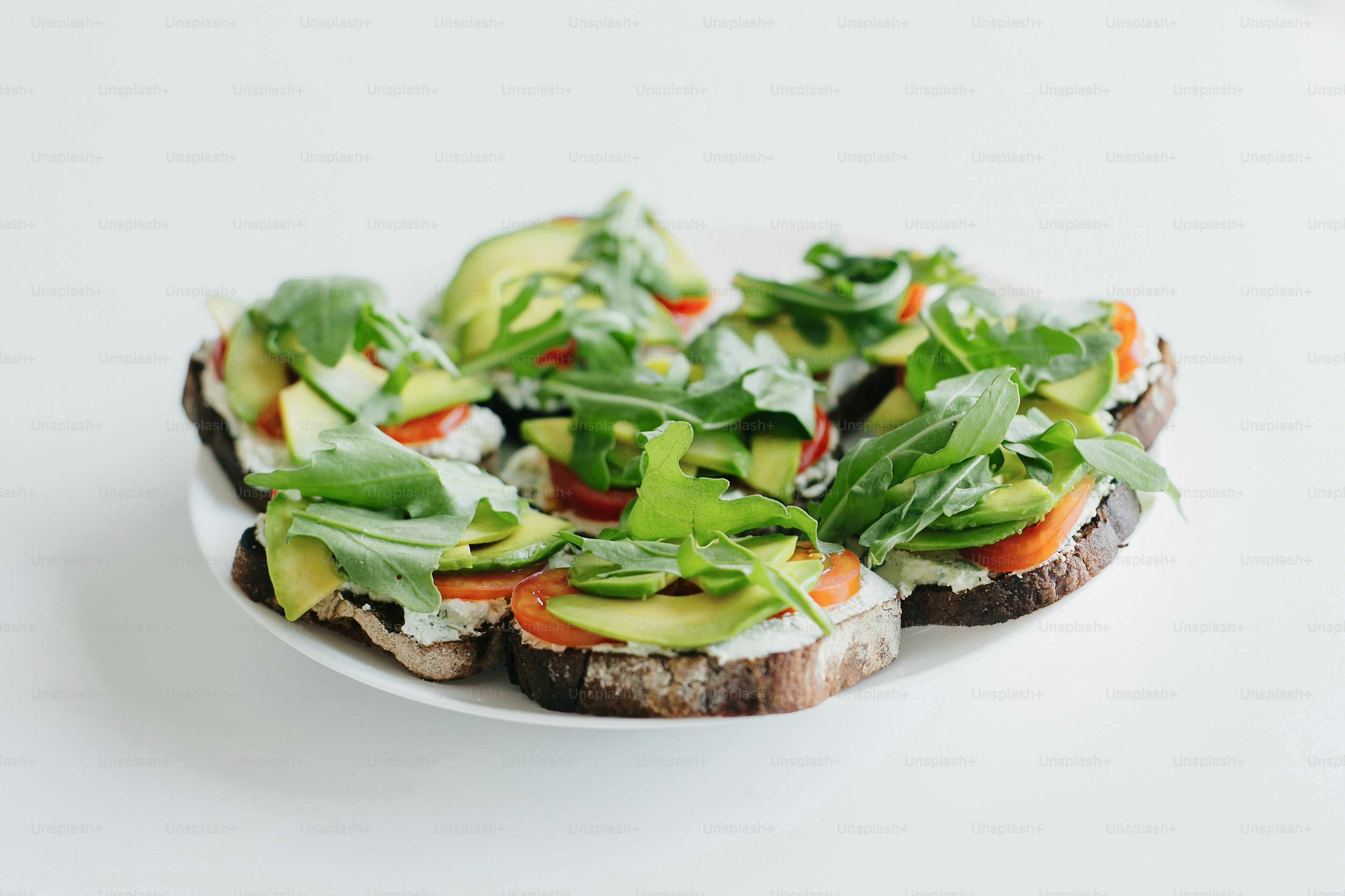 Homemade avocado toast on plate on white countertop in modern kitchen. Sandwich made of whole grain bread, avocado, tomato, arugula and cheese. Healthy eating and Home cooking concept.