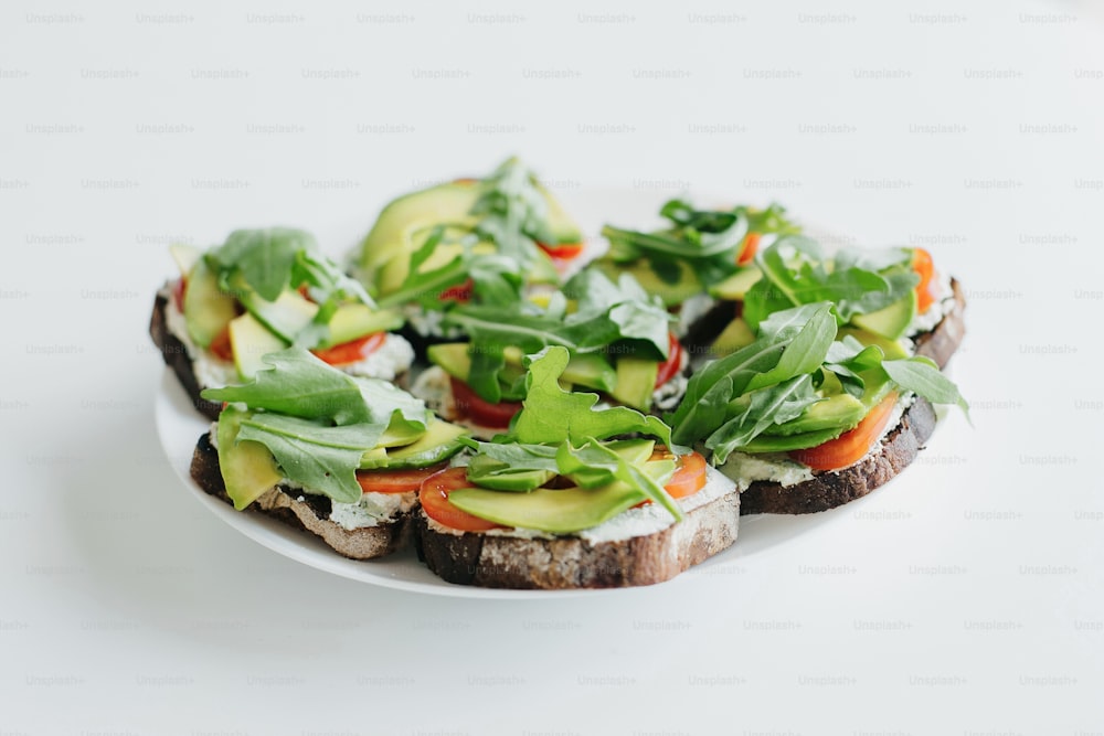 Hausgemachter Avocado-Toast auf dem Teller auf weißer Arbeitsplatte in moderner Küche. Sandwich aus Vollkornbrot, Avocado, Tomaten, Rucola und Käse. Gesunde Ernährung und Hausmannskost-Konzept.