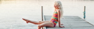 재미있고 행복한 귀여운 백인 금발 소녀가 호수 옆 나무 갑판 부두에 앉아 있다. 수영복을 입고 웃고 있는 아이가 물에 다리를 담그고 튀고 있다. 여름의 재미있는 야외 활동. 웹 배너 헤더.